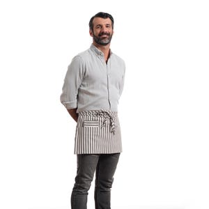 Puede incluir: Un hombre con una camisa de botones gris claro y un delantal a rayas. Est&aacute; sonriendo y mirando a la c&aacute;mara.