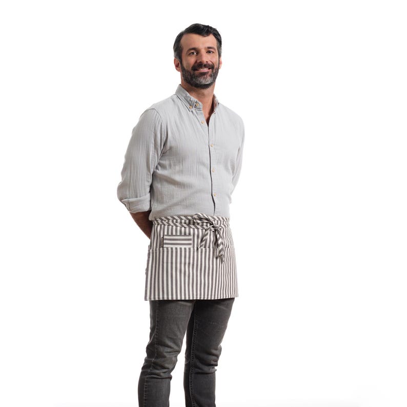 Puede incluir: Un hombre con una camisa de botones gris claro y un delantal a rayas. Est&aacute; sonriendo y mirando a la c&aacute;mara.