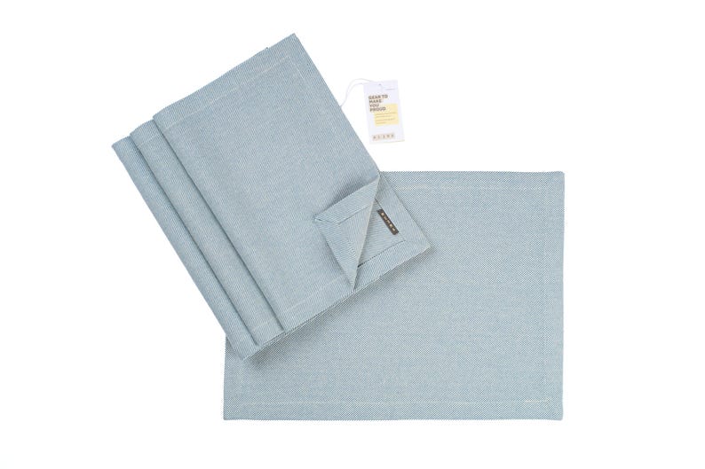 May include: Set of four light blue placemats made of woven fabric. The placemats are folded and stacked on top of each other. The placemats have a subtle texture and a slightly frayed edge.