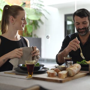 May include: A couple sits at a table with a wooden cutting board, a black plate with a salad, a glass of red wine, and a loaf of bread. The woman is holding a striped kitchen towel and the man is using a gold fork to eat his salad.