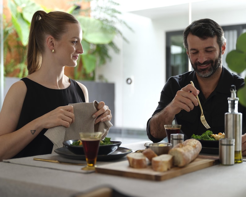 May include: A couple sits at a table with a wooden cutting board, a black plate with a salad, a glass of red wine, and a loaf of bread. The woman is holding a striped kitchen towel and the man is using a gold fork to eat his salad.