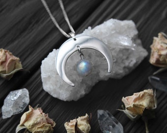 May include: A silver crescent moon pendant necklace with a dangling iridescent moonstone charm. The necklace is displayed on a white crystal cluster and surrounded by dried roses.