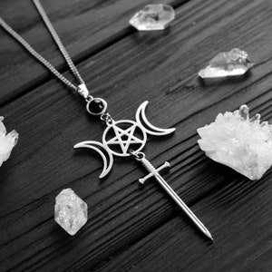 May include: A silver necklace with a pendant featuring a triple moon symbol, a pentagram, and a sword. The pendant is surrounded by clear quartz crystals on a dark wooden surface.