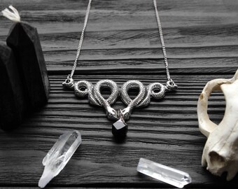 May include: A silver-toned necklace featuring two intertwined snakes, each with detailed scales, holding a black gemstone. The necklace is displayed on a dark wooden surface, alongside crystals, black candles, and a skull.