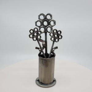 May include: A metal sculpture of a flower arrangement made from nuts and bolts. The sculpture is in a silver vase with a textured surface.