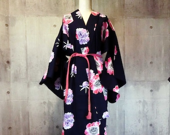 Puede incluir: Un kimono azul marino con un estampado floral en rosa y morado. El kimono está atado con un cinturón rosa. La prenda se exhibe en un maniquí contra una pared de hormigón gris.