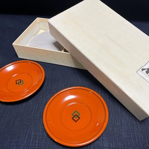 以下が含まれることがあります： 中央に幾何学模様が施された、オレンジ色の漆塗りの丸い小皿が2つ。小皿は濃い青色の表面に置かれている。背景には、日本語の文字が書かれた白い箱がある。