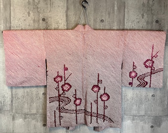 Könnte beinhalten: Ein rosa-weißes japanisches Kimono mit Blumenmuster. Das Kimono hängt an einem Kleiderbügel an einer Betonwand.