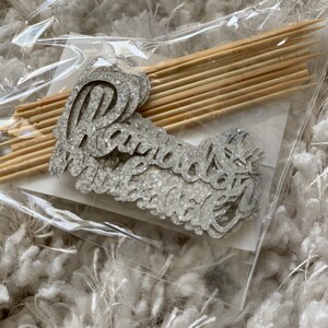 May include: A package of silver glitter cake toppers with the word "radiant" on them. The toppers are packaged with wooden skewers.