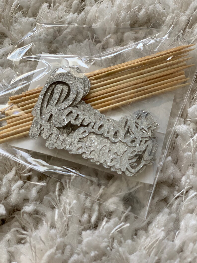 May include: A package of silver glitter cake toppers with the word "radiant" on them. The toppers are packaged with wooden skewers.