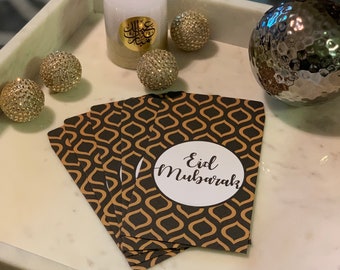 May include: A stack of gold and black patterned gift envelopes with the text "Eid Mubarak" on a white circle. The envelopes are on a white marble surface with a white candle and gold decorative balls.