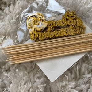 May include: A set of gold glitter cupcake toppers with the word "Congratulations" on them. The toppers are packaged with wooden skewers.