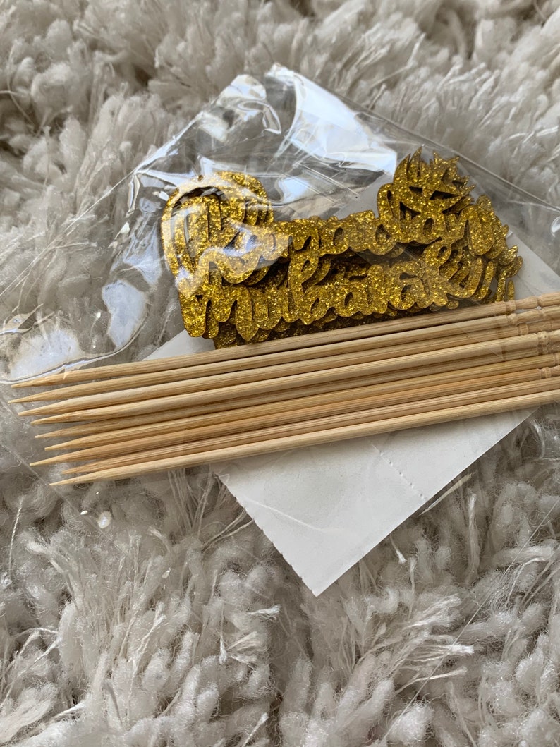 May include: A set of gold glitter cupcake toppers with the word "Congratulations" on them. The toppers are packaged with wooden skewers.