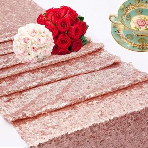 Op de afbeelding: Een roze gouden pailletten tafelloper met een boeket rode rozen en een boeket witte en roze rozen. Een vintage kop en schotel met een bloemenontwerp staan ook op de afbeelding. De tafelloper is een decoratief item.
