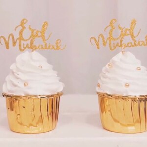 May include: Two gold cupcake liners with white frosting and gold glitter cupcake toppers that say "Eid Mubarak".