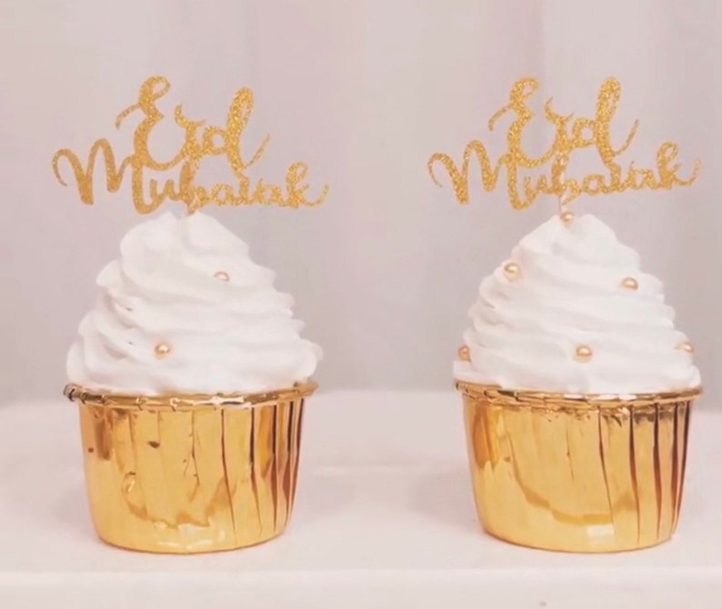 May include: Two gold cupcake liners with white frosting and gold glitter cupcake toppers that say "Eid Mubarak".