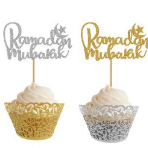 May include: Two cupcake toppers with the words "Ramadan Mubarak" in silver glitter and gold glitter. The toppers are on top of cupcakes with gold and silver cupcake liners.