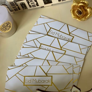 Könnte beinhalten: Ein Stapel weißer Umschläge mit goldenen geometrischen Mustern und dem aufgedruckten Text "Eid Mubarak!" Die Umschläge liegen auf einer hellbraunen Oberfläche.