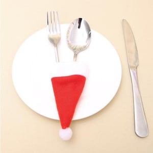 May include: A white plate with a fork and spoon wrapped in a red and white Santa hat. A silver knife is on the right side of the plate.