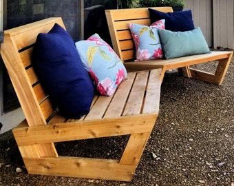 May include: Two wooden outdoor chairs with a natural wood finish. The chairs have slatted backs and seats. The chairs are decorated with floral patterned throw pillows in shades of blue, pink, and green.