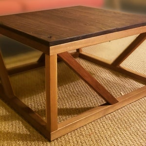 Puede incluir: Una mesa de centro de madera marrón oscuro con una base geométrica angulada. La mesa tiene una tapa rectangular y un marco robusto y abierto.