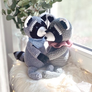 Könnte beinhalten: Zwei gehäkelte Waschbär-Stofftiere in Grau, die sich an den Händen halten und küssen. Jeder Waschbär hat eine schwarze Maske, ein weißes Gesicht und einen Schal. Einer trägt einen blauen Schal, der andere einen rosa Schal. Sie sitzen auf einer weißen, flauschigen Oberfläche in der Nähe eines Fensters.