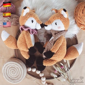 May include: Two crocheted fox plushies with brown and white fur, black eyes, and pink noses. One fox is wearing a pink ruffled collar and the other is wearing a brown scarf. The plushies are sitting on a wooden tray with a white background.