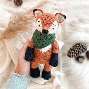 Può includere: Un giocattolo di peluche di volpe all'uncinetto con una sciarpa verde. La volpe ha una pelliccia arancione, occhi neri e un naso nero.