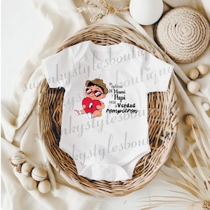 May include: White baby onesie with a cartoon graphic of a baby wearing sunglasses and a hat, holding a red heart. Text on the onesie reads "Un aplause pa' Mami y Papi porque en Verdad rompieron."