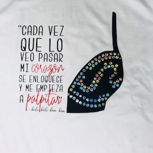May include: White t-shirt with black text that reads "CADA VEZ QUE LO VEO PASAR MI corazon SE ENLOQUECE Y ME EMPIEZA A palpitar" and a black silhouette of a high heel shoe with iridescent rhinestones. The text below the quote reads "bule bule bom bom"