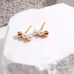 Könnte beinhalten: Ein Paar goldfarbene Ohrstecker. Jeder Ohrring hat eine kleine goldene Kugel, zwei weiße Perlen und einen goldenen Stift. Die Ohrringe werden auf einer weißen, strukturierten Oberfläche präsentiert.