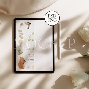Op de afbeelding: Een digitale tablet-mockup met een zwart frame, met een ontwerp voor een babyborreluitnodiging met aquarel illustraties van een beer, vogel en bloemen. Een witte stylus ligt naast de tablet, met witte tulpen op de achtergrond.