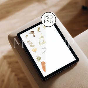 Può includere: Un tablet nero che mostra un invito a un baby shower con illustrazioni ad acquerello di un uccello, fiori e una volpe. Il testo dell'invito dice "oh baby". Il tablet è appoggiato su una superficie imbottita beige.