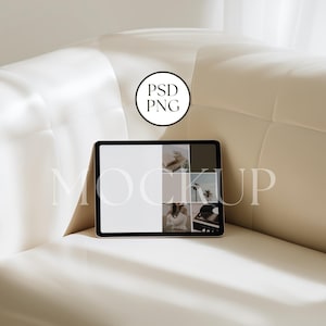 May include: A black tablet with a white screen and a collage of images is displayed on a cream-coloured sofa. The tablet is positioned in the centre of the frame, with the text "MOCKUP" in a light colour across the bottom of the screen.