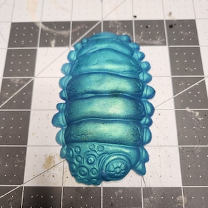 Puede incluir: Una criatura segmentada de color azul turquesa, impresa en 3D, con un patrón circular detallado en la parte inferior.