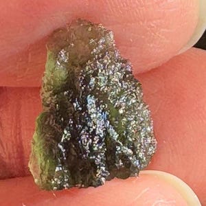 May include: A small, irregular piece of translucent olive-green moldavite with a textured, pitted surface. The stone displays a deep green hue with areas of light reflection and a jagged, natural edge, showcasing its unique geological formation.