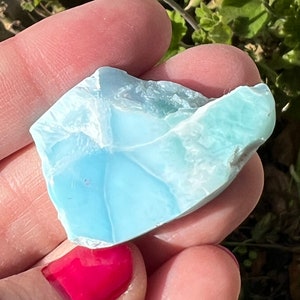 Puede incluir: Una pieza de piedra Larimar de forma irregular, translúcida y azul claro. La piedra tiene una superficie lisa y pulida y se sostiene en una mano.