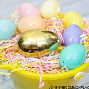 May include: A yellow bucket filled with colorful Easter eggs and shredded paper. The eggs are pastel shades of pink, blue, yellow, and purple, with one gold egg in the center. The shredded paper is pink, yellow, and white.
