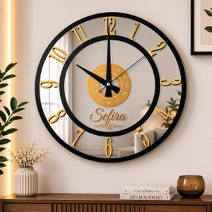 Peut inclure: Une horloge murale ronde avec un cadre noir et un cadran miroir. Elle présente des chiffres dorés de 1 à 12, des aiguilles noires et un détail circulaire doré au centre avec de petits chiffres. Le nom Sefira est inscrit en lettres dorées.