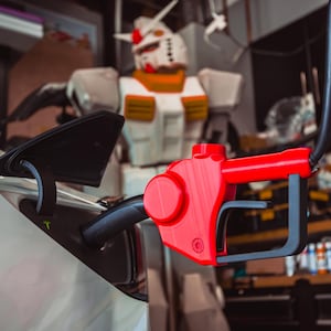 Pode incluir: Um bico de bomba de gasolina vermelho e preto com uma mangueira preta conectada a um carro elétrico branco. O carro está carregando em uma garagem com um grande robô ao fundo.