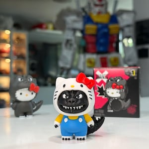 Puede incluir: Una figura de Hello Kitty vestida con un disfraz de Godzilla negro con un lazo rojo, mangas amarillas y peto azul. La figura presenta una cabeza de gato blanca, dientes afilados y una cola negra. Se observa una caja a juego y otra figura cerca.