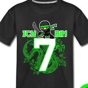 Op de afbeelding: Zwart T-shirt met een groene draak en een ninja die een zwaard vasthoudt. Het getal 7 is in wit met de tekst "Ich bin" in groen. De tekst "F&auml;llt kleiner aus. Bitte gr&ouml;&szlig;en-Tabelle beachten." is in groen op een witte cirkel.