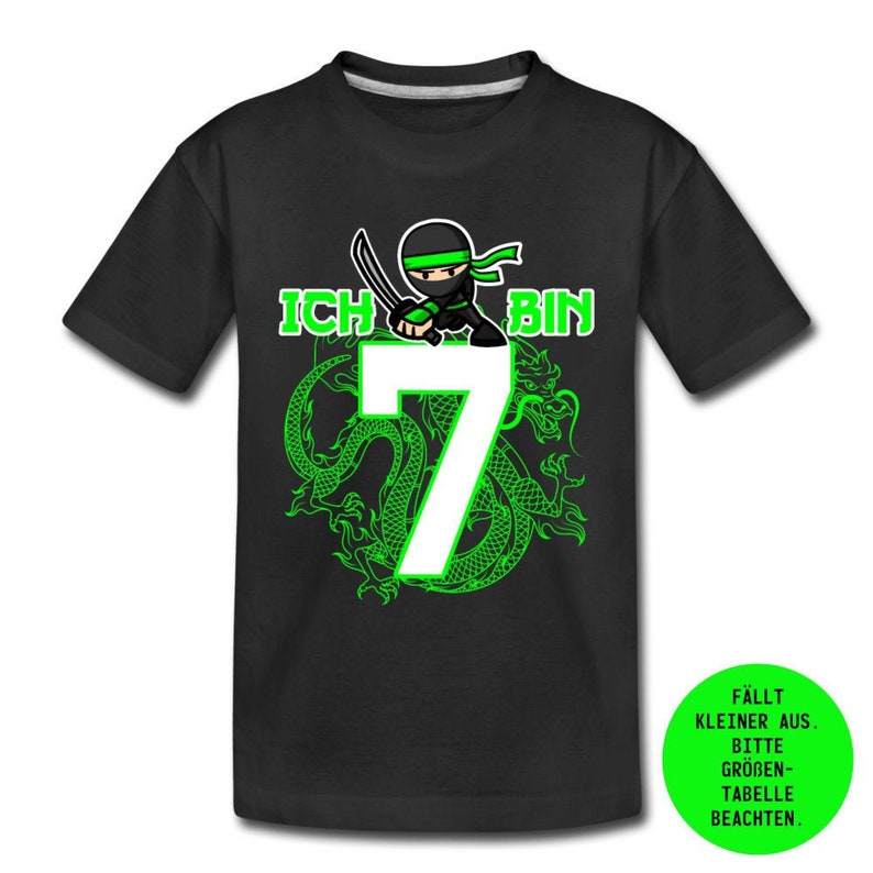 Op de afbeelding: Zwart T-shirt met een groene draak en een ninja die een zwaard vasthoudt. Het getal 7 is in wit met de tekst "Ich bin" in groen. De tekst "F&auml;llt kleiner aus. Bitte gr&ouml;&szlig;en-Tabelle beachten." is in groen op een witte cirkel.