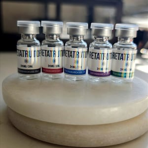 May include: Five clear glass vials with silver caps, each containing a liquid. The labels read RETATRUTIDE, 24MG/3ML, and FOR RESEARCH USE ONLY. Each label features a unique colour accent: black, red, blue, purple, and green, with a small atom icon.