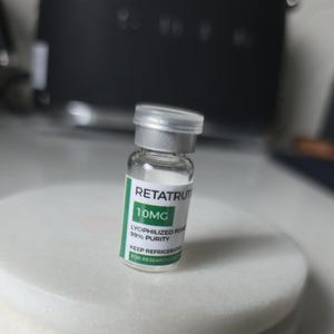 Peut inclure: Un petit flacon en verre transparent avec un bouchon en métal argenté et une étiquette blanche à accents verts. Le texte indique RETATRUT, 10MG, LYOPHILIZED POWDER, 99% PURITY, KEEP REFRIGERATED et FOR RESEARCH USE ONLY. Le flacon contient une poudre.