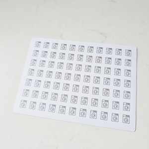 May include: A sheet of white stickers featuring a repeating pattern of small, black line-drawn washing machine icons. The stickers are rectangular with rounded corners and are arranged in a grid pattern.
