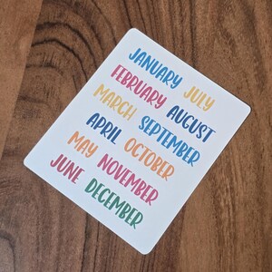 Pode incluir: Um cartão retangular branco com os meses do ano escritos em letras coloridas em estilo manuscrito. Os meses estão dispostos em duas colunas, com janeiro a junho à esquerda e julho a dezembro à direita.