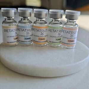 Peut inclure: Cinq flacons en verre transparent avec des bouchons argentés, contenant chacun une substance blanche. Les étiquettes indiquent RETATRUTIDE, LYOPHILIZED PEPTIDE, 10MG, FOR RESEARCH PURPOSES ONLY, avec des bandes colorées jaune, bleue, orange, verte et rose.