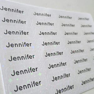 May include: A sheet of rectangular adhesive labels featuring the name Jennifer printed in a bold black sans-serif font. Each individual label has a holographic finish that displays subtle star-shaped light reflections across the surface.