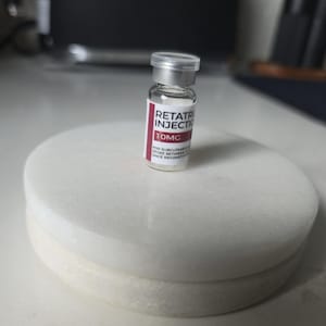 Peut inclure: Un petit flacon en verre transparent avec un bouchon argenté et une étiquette blanche portant le texte rouge RETATRU INJECTION 10MG, ainsi que des instructions pour l'utilisation sous-cutanée et les conditions de stockage.